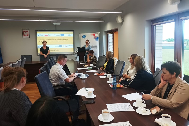 Na pierwszym planie uczestnicy szkolenia siedzą po obu stronach stołu i patrzą w kierunku prowadzących. W tle prezentacja i pracownicy biura LGD.