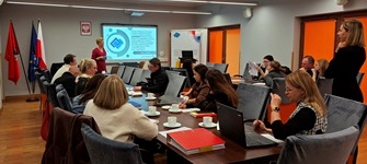 Uczestnicy szkolenia siedzą przy stołach i robią notatki. Głowami są zwróceni na prowadzące.